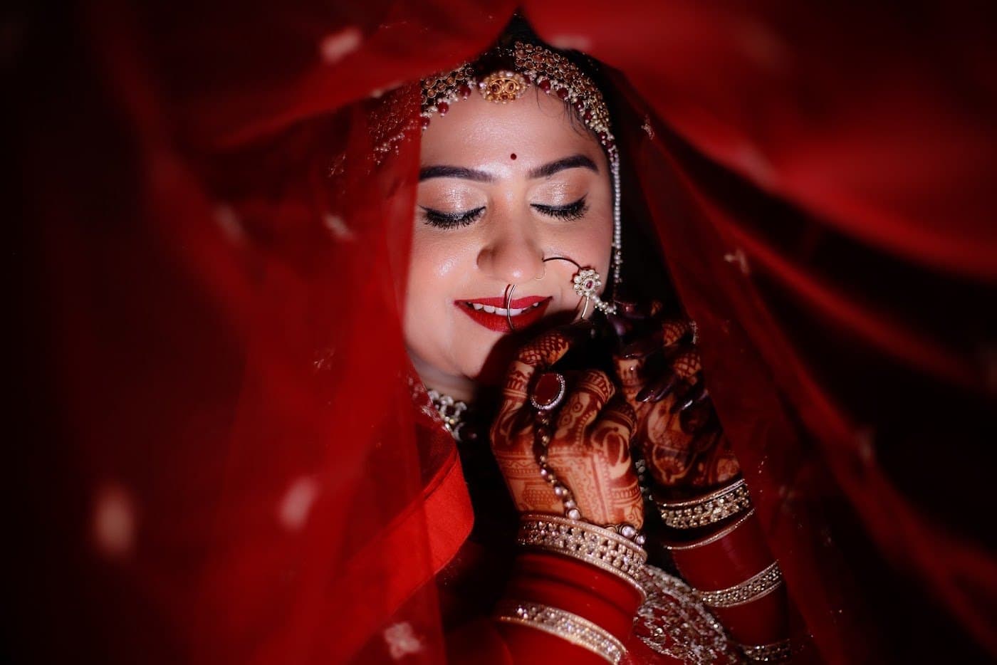 Smiling bride under a red veil with luxury bridal makeup by Anuja Paul Makeup Studio, Panna
